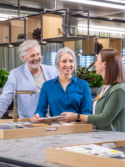 Drei Personen stehen an einem Küchenausstellungsbereich. Eine Frau zeigt einem Paar verschiedene Materialproben oder Farbmuster. Drei Personen stehen an einem Küchenausstellungsbereich. Eine Frau zeigt einem Paar verschiedene Materialproben oder Farbmuster.