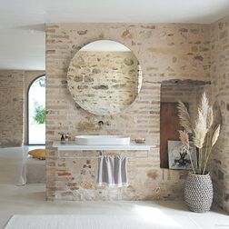 Ein Badezimmer mit rustikaler Backsteinwand und rundem Spiegel. Ein moderner Waschtisch, Dekorationen wie ein Korb mit Pampasgras und ein helles, einladendes Ambiente runden den Raum ab. Ein Badezimmer mit rustikaler Backsteinwand und rundem Spiegel. Ein moderner Waschtisch, Dekorationen wie ein Korb mit Pampasgras und ein helles, einladendes Ambiente runden den Raum ab.